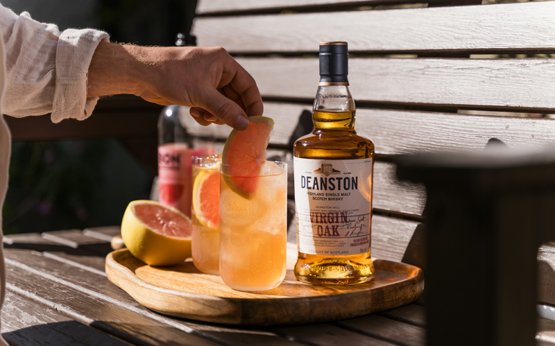 Pink Grapefruit Spritz being prepared on a garden bench with a bottle of Deanston Virgin Oak