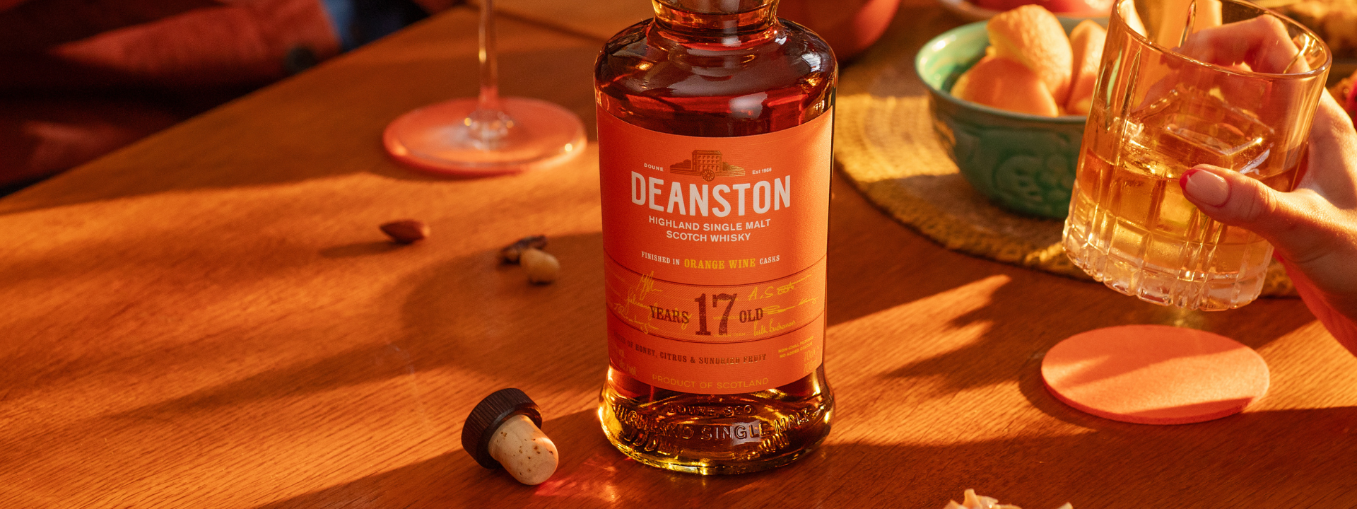Deanston whisky bottle on a wooden table with a glass of whisky held by a hand.