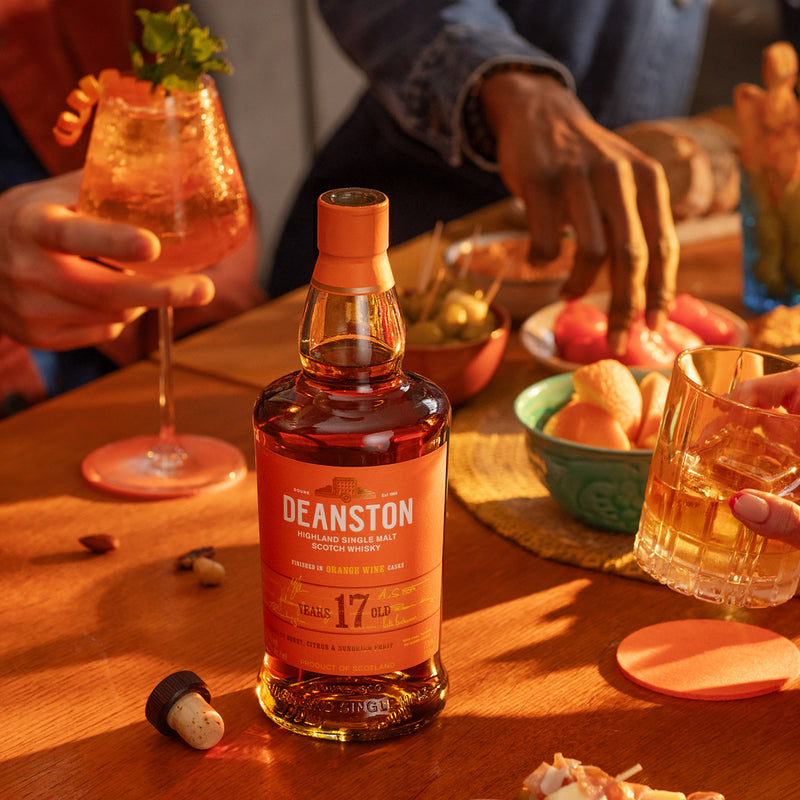 An open bottle of Deanston 17 Year Old Orange Wine Cask Finish Single Malt is on a wooden table amongst bowls of food suggesting tapas. Three people are around the table; one is holding a Spritz cocktail, one is holding a lowball glass of whisky with a large ice cube and one is picking up an orange wedge from the spread of tapas.