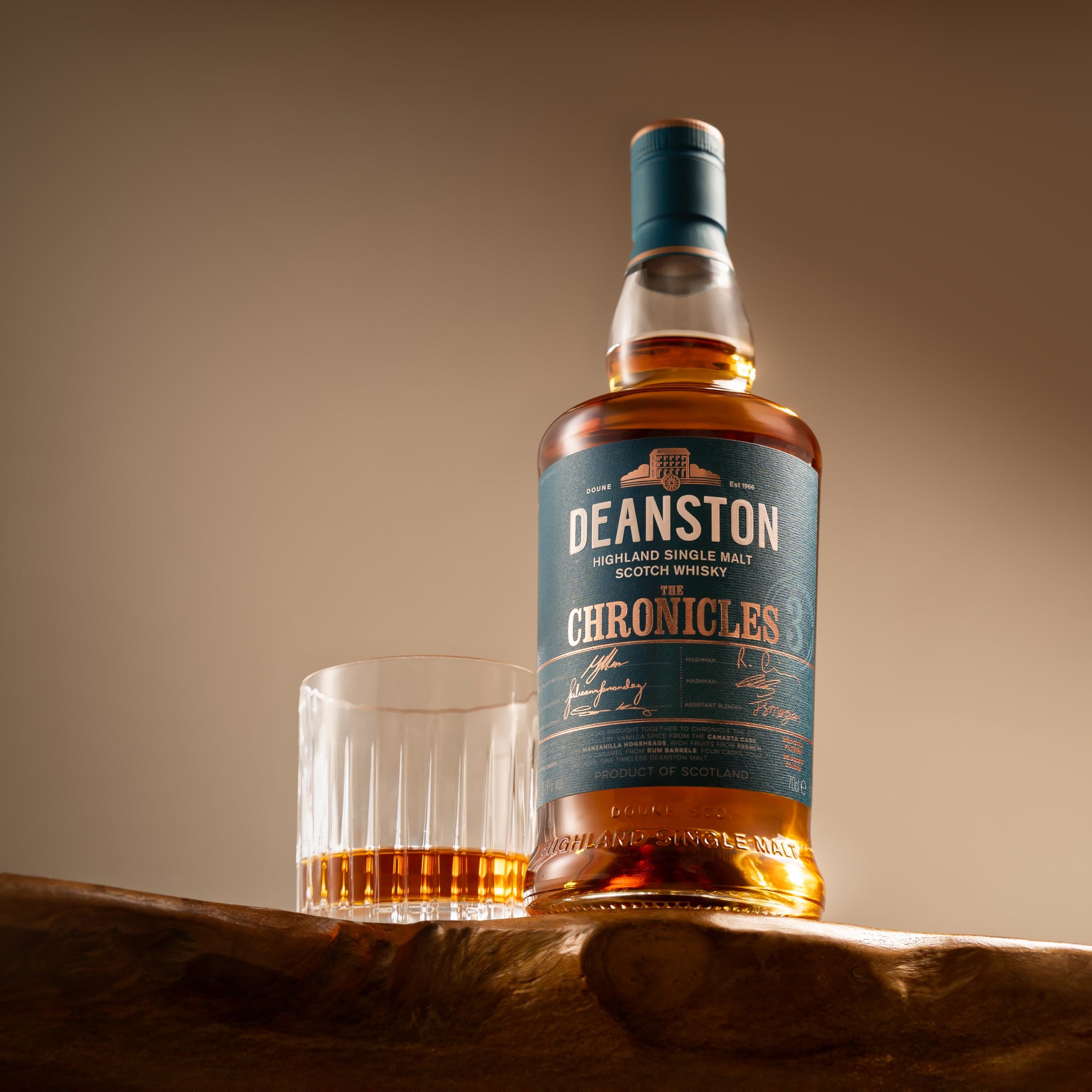 A bottle of Deanston Chronicles Series: Edition 3 whisky next to a filled glass on a wooden surface, with the label detailing the product name and distillery information.