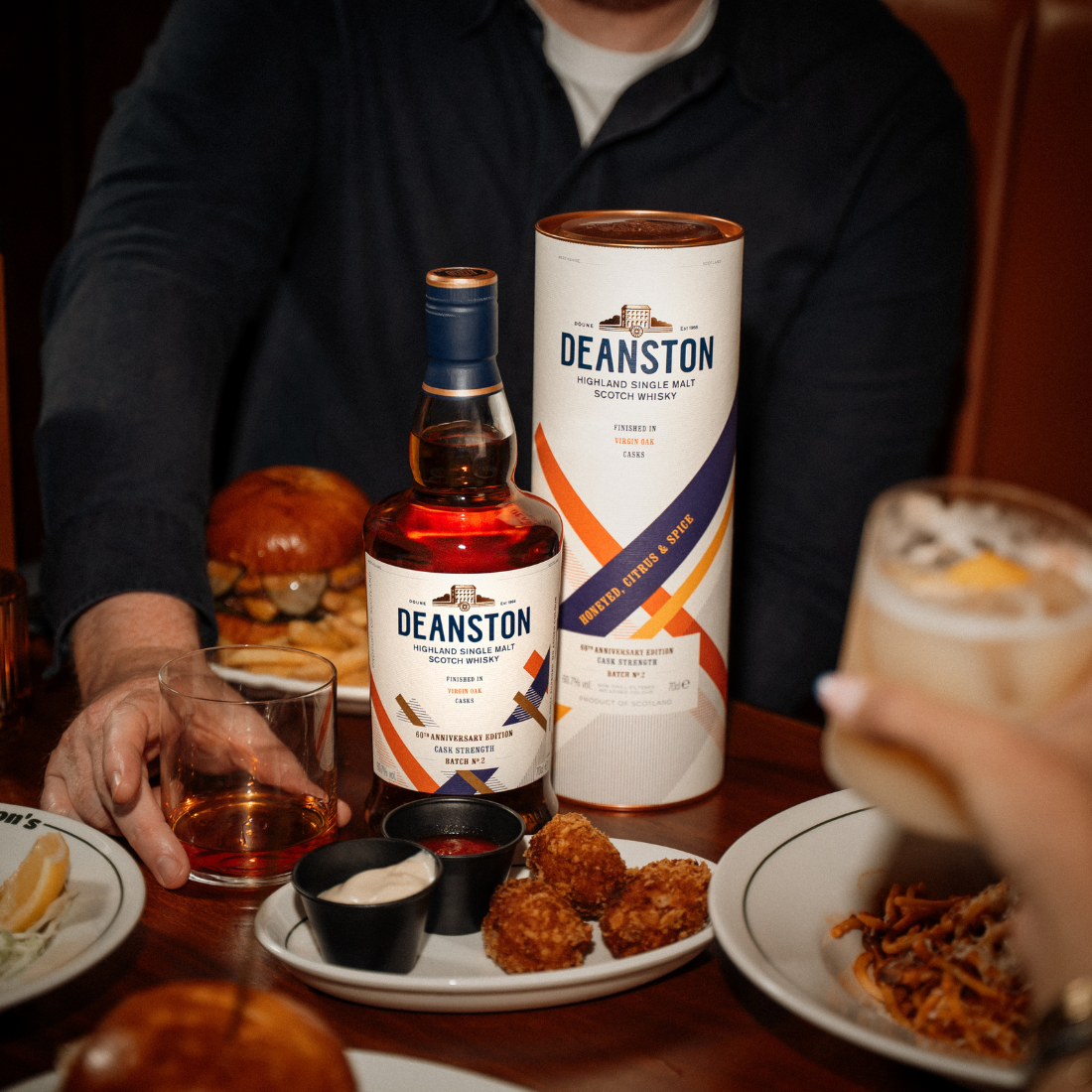 A bottle of Virgin Oak Cask Strength: 2026 Edition is sitting on a brown wooden table next to its matching pack. The table is fully of food and two people are sitting opposite each other.