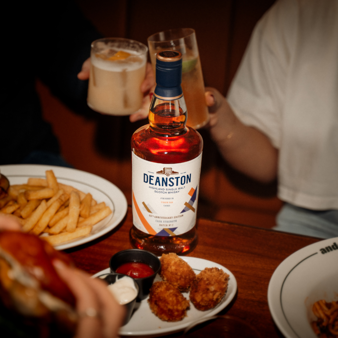 Three people are sitting around a table with food and cocktails. A bottle of Virgin Oak Cask Strength: 2026 Edition is in the middle of the table.