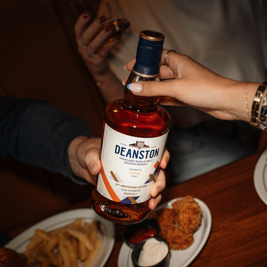 A botle of Virgin Oak Cask Strength:2026 Edition is being passed between two people across a table of food.